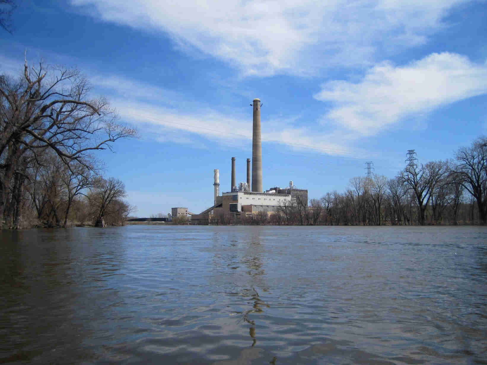 View from the middle of a river with bare trees on the sides of it, and a building with a smoke stack ahead